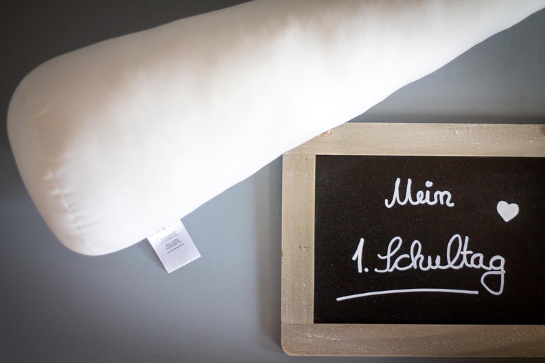 May include: A white pillow with a tag on it is next to a small chalkboard with the words "Mein 1. Schultag" written in white chalk.