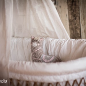 May include: A gray and pink llama stuffed animal sits in a white wicker bassinet with a white sheet. The bassinet is surrounded by a white fabric canopy.