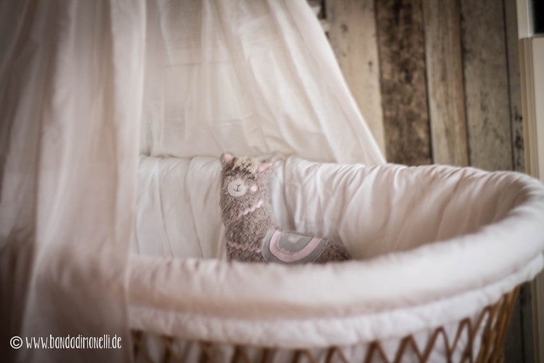 May include: A gray and pink llama stuffed animal sits in a white wicker bassinet with a white sheet. The bassinet is surrounded by a white fabric canopy.