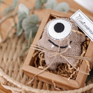 May include: A brown plush monster toy with a single large eye and a toothy grin, packaged in a brown cardboard box with shredded paper and a tag that reads "Mutmonster".