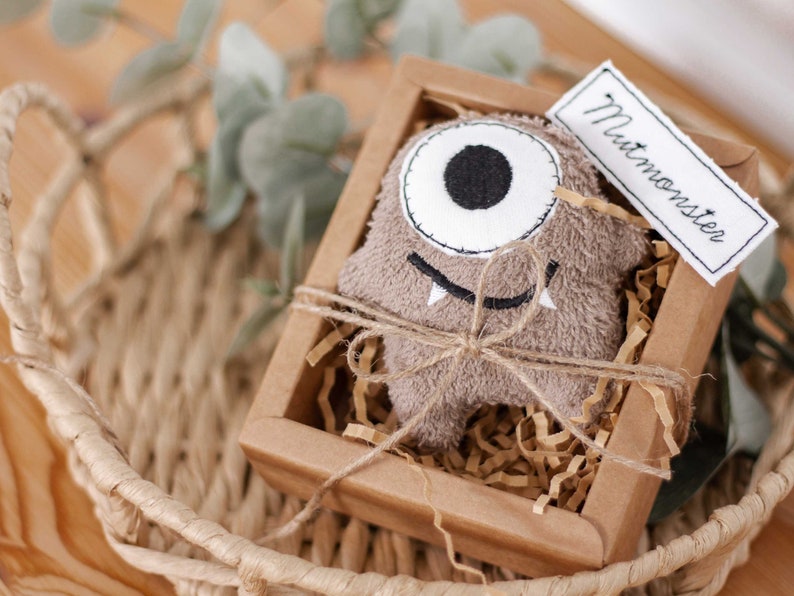 May include: A brown plush monster toy with a single large eye and a toothy grin, packaged in a brown cardboard box with shredded paper and a tag that reads "Mutmonster".