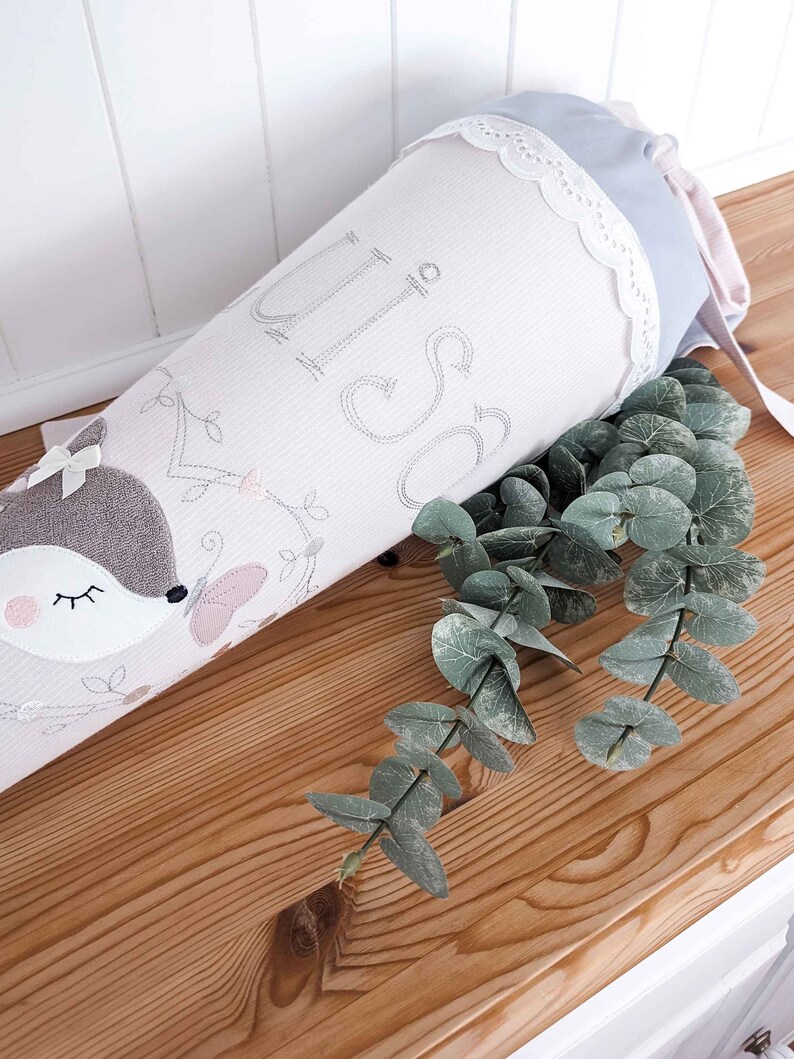 May include: A light pink and gray fabric cone with a white lace trim and a cute embroidered fox design. The cone has the word "Liso" embroidered on it.