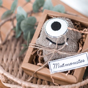 May include: A brown plush monster toy with one large eye and a toothy grin is packaged in a brown cardboard box with a white tag that reads "Mutmonster".