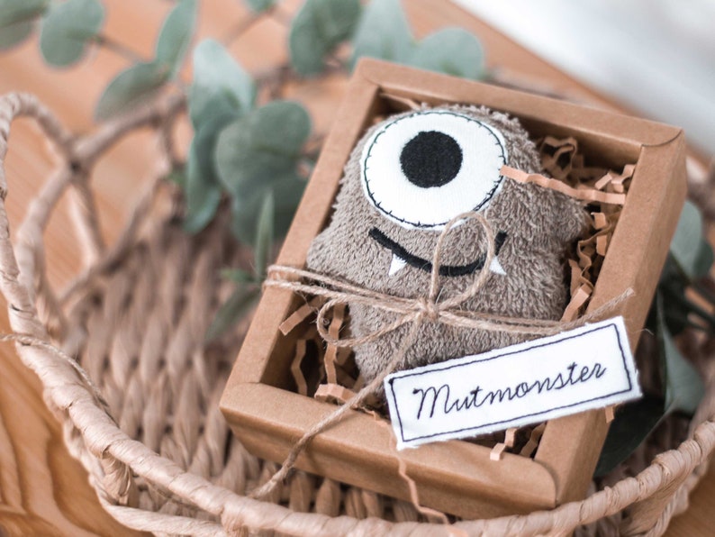 May include: A brown plush monster toy with one large eye and a toothy grin is packaged in a brown cardboard box with a white tag that reads "Mutmonster".