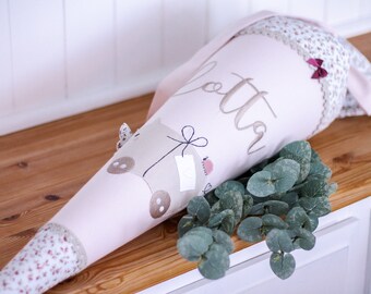 May include: A pink and white school cone with a floral pattern and a brown bear design. The cone is decorated with a pink bow and the name "Lotta" is embroidered on the side.