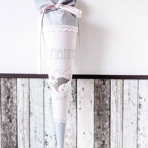 May include: A gray and pink fabric cone with white lace trim. The cone has a deer design and the name "Johanna" embroidered on it.