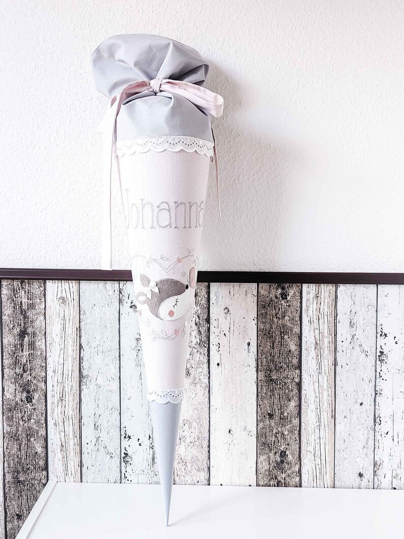 May include: A gray and pink fabric cone with white lace trim. The cone has a deer design and the name "Johanna" embroidered on it.