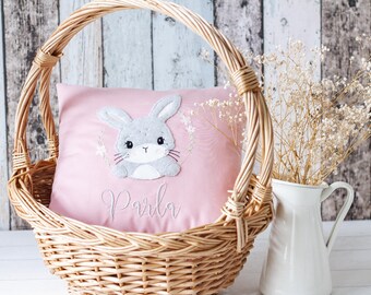 May include: A pink pillow with a gray embroidered bunny and the name "Parla" in white embroidery. The pillow is in a wicker basket.