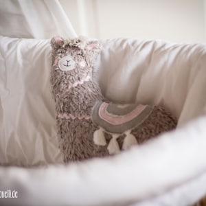 May include: A plush gray llama toy with pink accents and white tassels, sitting in a white wicker basket.