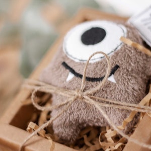 May include: A brown plush monster toy with a single white eye and a black mouth, tied with twine and placed in a brown cardboard box with shredded paper.