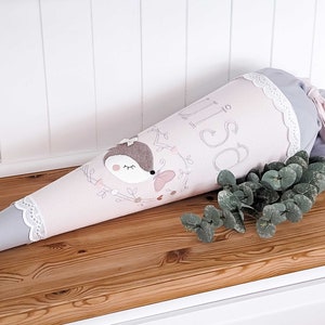 May include: A pink and gray school cone with a white lace trim. The cone has a cute embroidered hedgehog design and the name "Lisa" written on it.