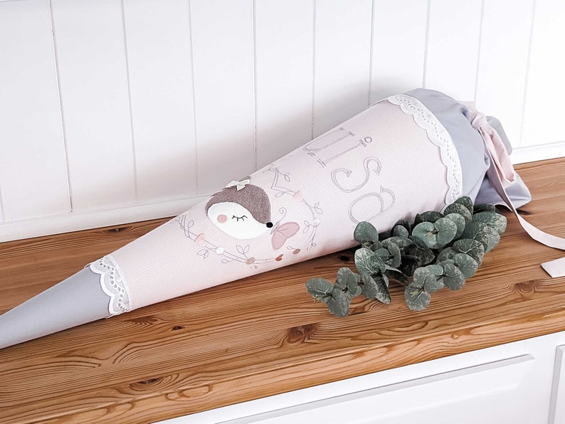 May include: A pink and gray school cone with a white lace trim. The cone has a cute embroidered hedgehog design and the name "Lisa" written on it.