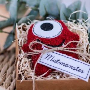 May include: A red plush monster with a white eye and a stitched mouth, tied with twine and a white tag that says "Mutmonster", in a brown box filled with shredded paper.