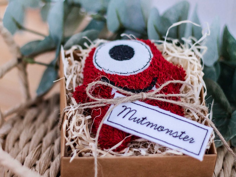 May include: A red plush monster with a white eye and a stitched mouth, tied with twine and a white tag that says "Mutmonster", in a brown box filled with shredded paper.