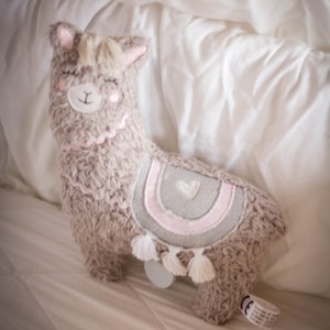 May include: A plush brown llama toy with a pink and gray heart design on its back. The llama has a white tassel on its tail and is lying on a white bed.
