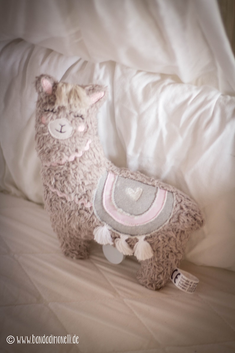 May include: A plush brown llama toy with a pink and gray heart design on its back. The llama has a white tassel on its tail and is lying on a white bed.