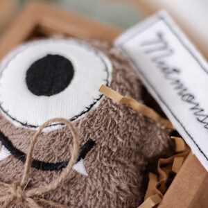 May include: A brown plush monster toy with a single white eye and a stitched-on mouth. The toy is tied with twine and has a white tag that says "The Monster".