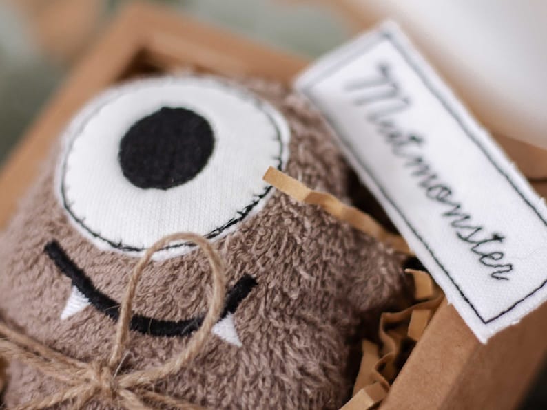 May include: A brown plush monster toy with a single white eye and a stitched-on mouth. The toy is tied with twine and has a white tag that says "The Monster".
