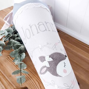 May include: A white and pink fabric cone with a gray deer face and a white bow. The cone has a white lace trim and the name "Johanna" is embroidered on the front.