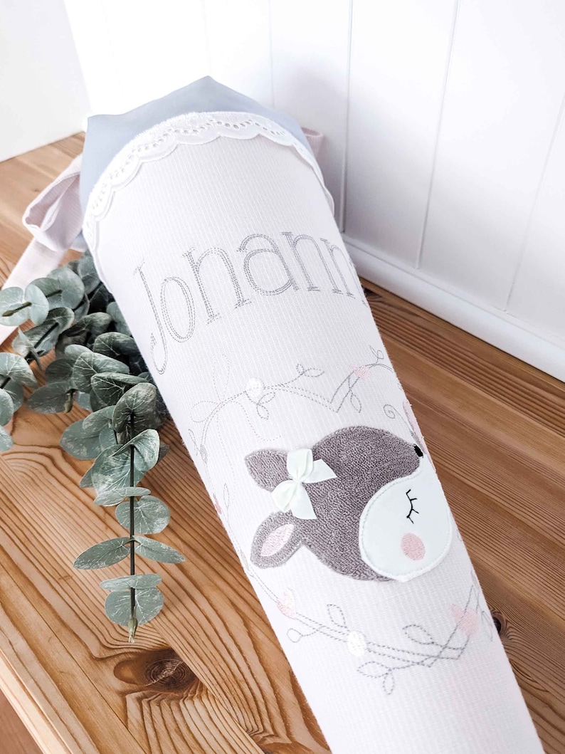 May include: A white and pink fabric cone with a gray deer face and a white bow. The cone has a white lace trim and the name "Johanna" is embroidered on the front.