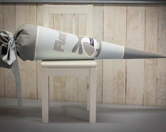 May include: A gray and light blue school cone with the name "Florian" embroidered on it. The cone is sitting on a white wooden chair.