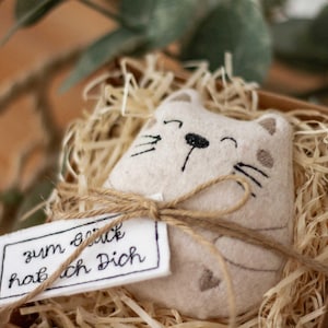 Könnte beinhalten: Ein beiges, katzenförmiges Plüschtier mit gestickten Gesichtszügen und einem herzförmigen Detail, eingebettet in ein Strohbett in einer kleinen Holzkiste. Ein Anhänger mit Text ist mit Bindfaden an dem Spielzeug befestigt.