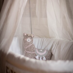 May include: A gray and pink llama plush toy sits in a white wicker bassinet with a white canopy. The llama has a pink heart on its back and white tassels.
