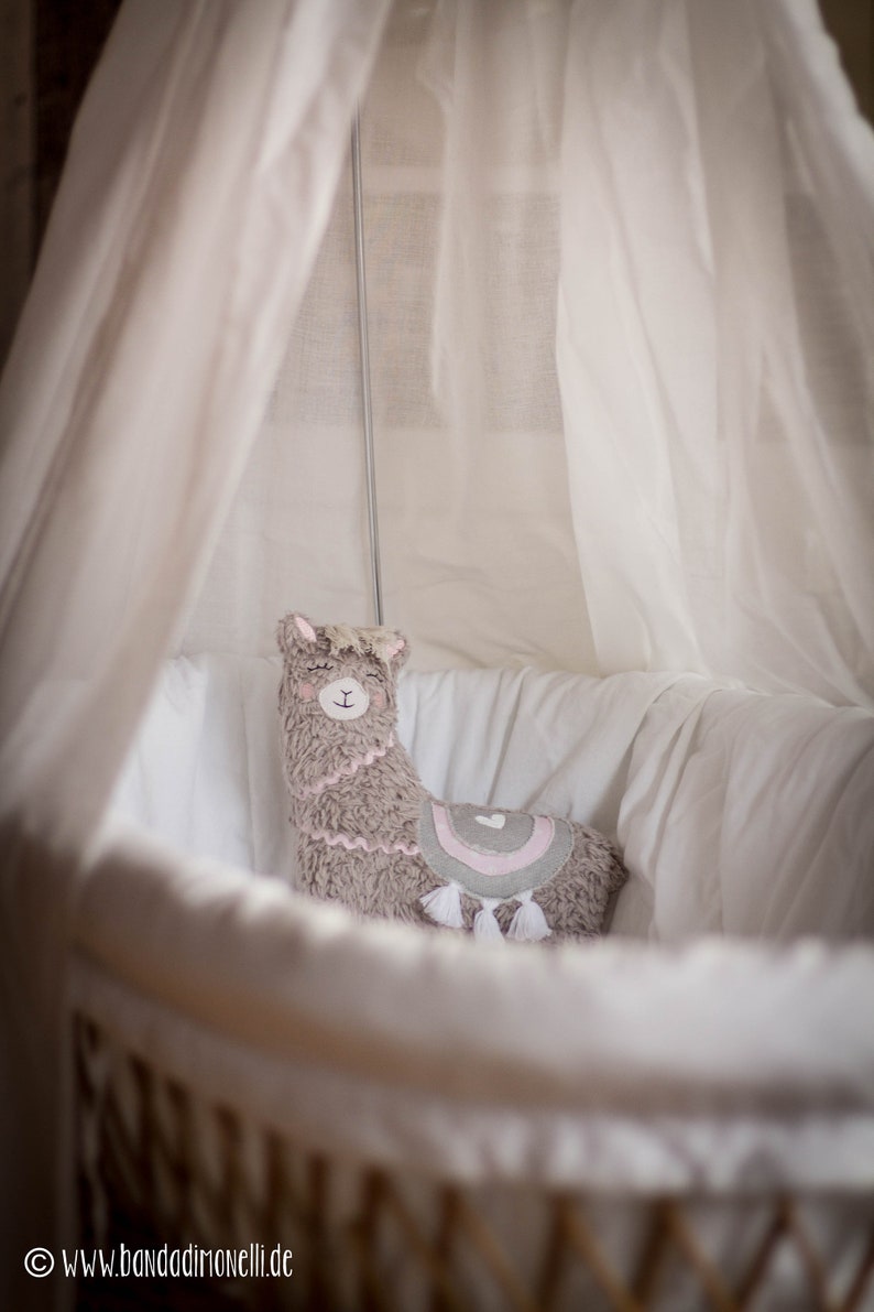 May include: A gray and pink llama plush toy sits in a white wicker bassinet with a white canopy. The llama has a pink heart on its back and white tassels.