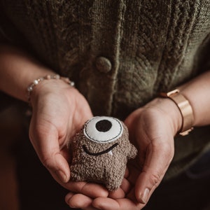 May include: A person is holding a brown plush toy with a single white eye and a smiling mouth. The toy is made of a soft, fuzzy material.