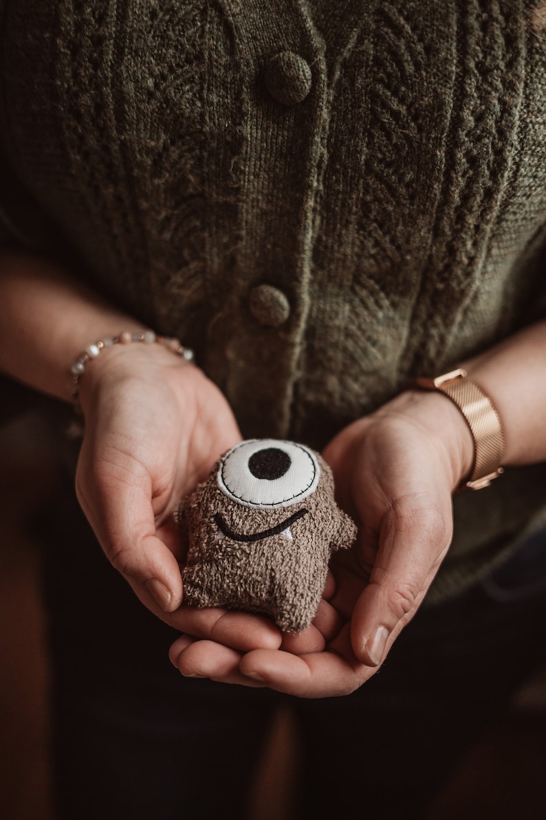 May include: A person is holding a brown plush toy with a single white eye and a smiling mouth. The toy is made of a soft, fuzzy material.