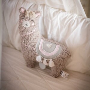 May include: A plush brown llama toy with a pink and gray striped design and white tassels. The llama has a white heart on its back and is lying on a white bed.