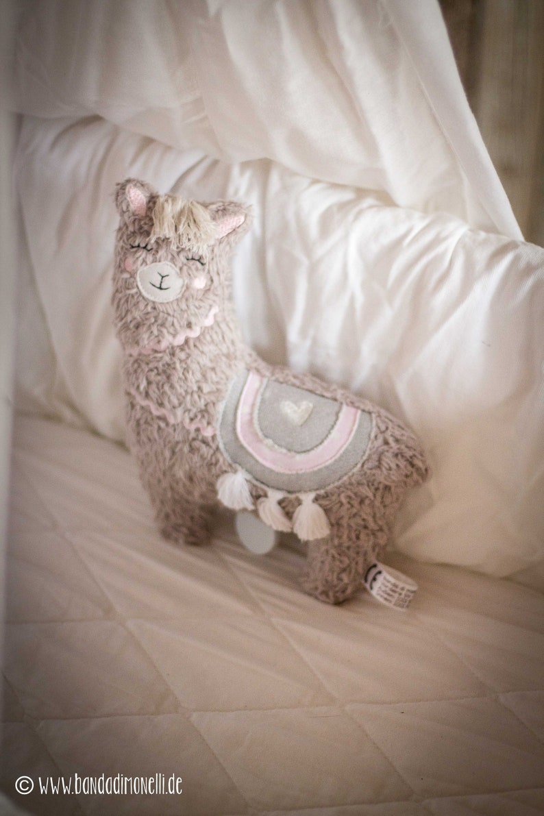 May include: A plush brown llama toy with a pink and gray striped design and white tassels. The llama has a white heart on its back and is lying on a white bed.