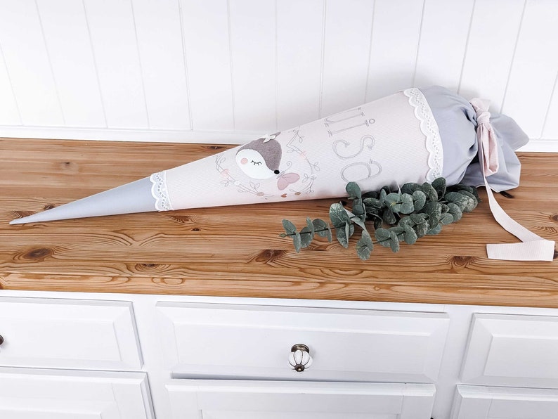 May include: A gray and pink fabric cone with a white lace trim and a pink ribbon. The cone has a deer design and the name "Isa" embroidered on it. The cone is sitting on a wooden surface with a sprig of eucalyptus.