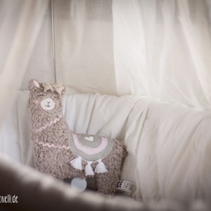 May include: A plush llama toy with a pink heart and rainbow design, sitting in a white crib with a white canopy.