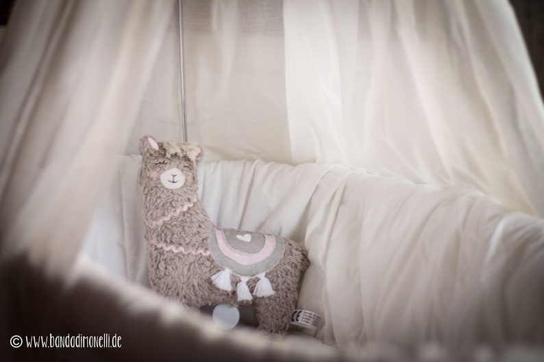 May include: A plush llama toy with a pink heart and rainbow design, sitting in a white crib with a white canopy.