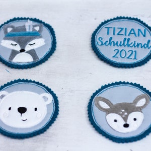 Könnte beinhalten: Vier runde Filzpatches mit blauem Rand. Ein Patch zeigt einen grauen Fuchs mit einem blauen Stirnband und Federn. Ein weiterer Patch zeigt einen weißen Eisbären. Ein dritter Patch zeigt ein graues Reh. Der letzte Patch ist hellgrau mit dem Text "TIZIAN Schulkinds 2021".