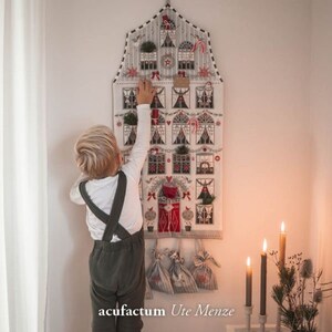Può includere: Un bambino raggiunge un calendario dell'avvento fatto a mano a forma di casa con 24 finestre numerate. Il calendario è decorato con dettagli rossi, bianchi e verdi, tra cui bastoncini di zucchero, ghirlande e piccoli alberi. Il calendario è appeso a un muro bianco. Il testo "acufractum Ute Menze" è visibile nella parte inferiore dell'immagine.