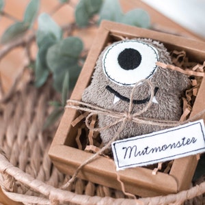 May include: A brown plush monster toy with one white eye and a stitched-on smile, packaged in a brown cardboard box with a white tag that reads "Mutmonster".