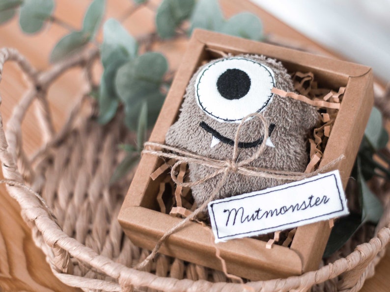 May include: A brown plush monster toy with one white eye and a stitched-on smile, packaged in a brown cardboard box with a white tag that reads "Mutmonster".