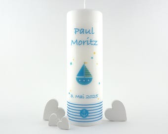 Peut inclure: Une bougie blanche ornée de décorations bleues sur le thème nautique. La bougie porte le nom "Paul Moritz" en bleu, un voilier bleu et la date "3. Mai 2025". Des rayures bleues et une ancre décorent la base. Plusieurs décorations blanches en forme de cœur sont devant la bougie.
