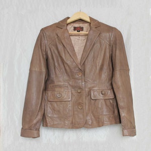 Puede incluir: Una chaqueta de cuero marrón con cierre de botones y dos bolsillos de solapa. La chaqueta tiene un cuello de muesca y mangas largas.