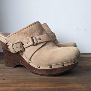 Könnte beinhalten: Beigefarbene Wildledersandalen mit Keilabsatz und Schnalle. Die Sandalen haben eine Holzplattform und einen dicken Keilabsatz.