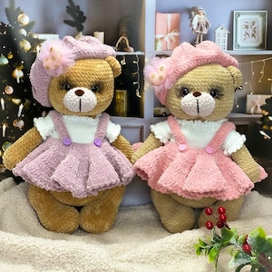 Puede incluir: Dos osos de peluche tejidos a crochet que llevan vestidos y sombreros rosas con lazos. Los osos están sentados sobre una superficie blanca y esponjosa con una planta verde con bayas rojas en primer plano.