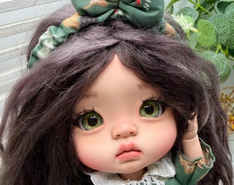 Op de afbeelding: Een pop met donkerbruin haar draagt een groene haarband met een grote stoffen strik en een groene jurk met een witte kanten kraag. De jurk en haarband zijn versierd met een patroon van bruine bladeren, groen loof en kleine witte bloemen.