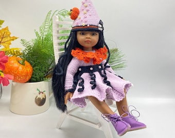 May include: A doll wearing a purple crocheted dress with black and orange accents, a purple and white crocheted hat with beads, and purple shoes. The doll is sitting on a white wooden chair.