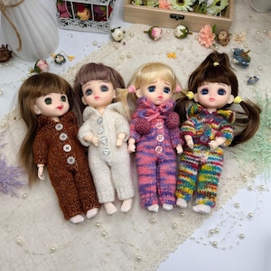 May include: Four small dolls wearing knitted hooded jumpsuits in various colors. The jumpsuits are brown, cream, pink and purple striped, and rainbow striped. Each doll has a different hairstyle and button details on the jumpsuit.