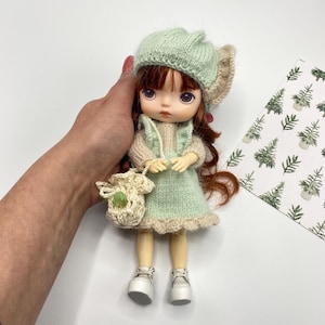Puede incluir: Una muñeca pequeña con un vestido de punto verde, un gorro de punto verde con una solapa de oreja beige y zapatos blancos. La muñeca lleva una pequeña bolsa de ganchillo.
