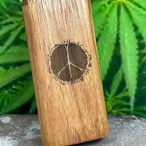 May include: A rectangular wooden box with a hinged lid. The box is light brown with a darker brown peace sign carved into the front. The background is a blurred green.