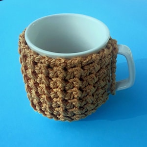 Könnte beinhalten: Eine weiße Keramiktasse mit einer gehäkelten Tasse in einem warmen Senfgelb. Die Tasse ist mit einem einfachen Stichmuster gearbeitet und hat eine lange Franse.