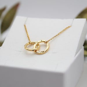 Könnte beinhalten: Eine goldene Halskette mit einer zarten Kette und einem Anhänger mit zwei ineinander verschlungenen Kreisen. Die Kreise sind strukturiert und haben eine gehämmerte Oberfläche.